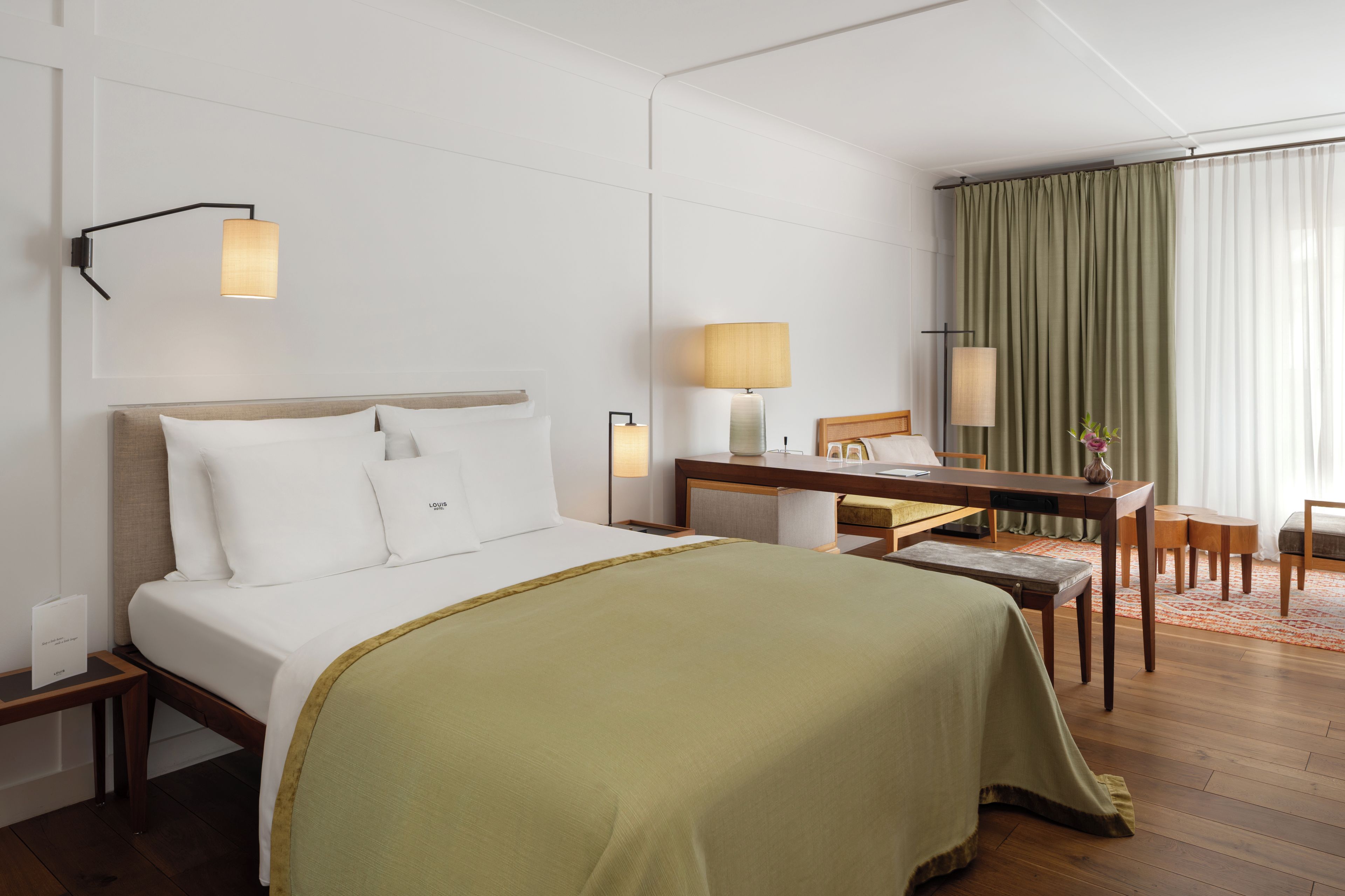 A hotel room with a bed, bedside table, desk, chair, and a view of a curtain.