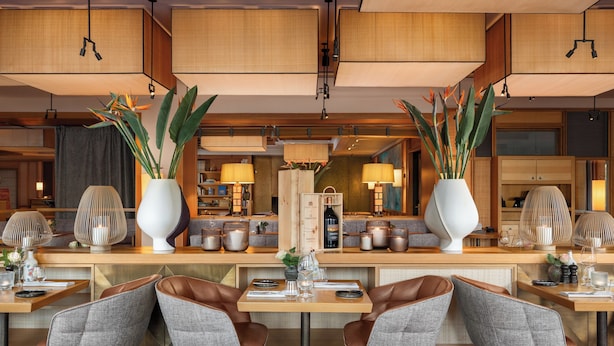 Un restaurante moderno con barra central, asientos con sillas acolchadas y jarrones decorativos con plantas.