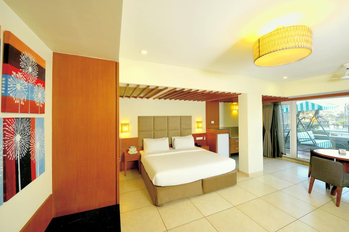 Habitación de hotel con cama, mesitas de noche, escritorio y una silla. Se ve una ventana con cortinas, una lámpara en el techo y una obra de arte en la pared.