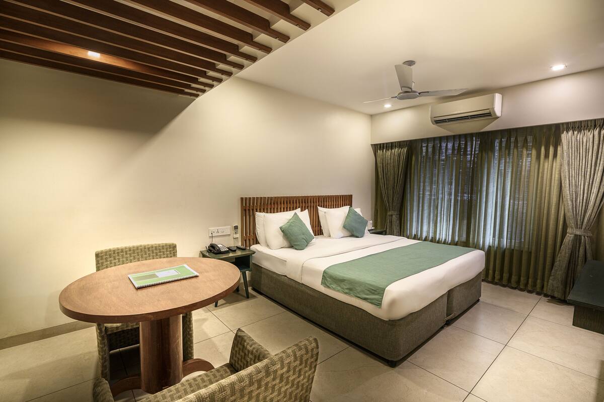 Habitación de hotel con una cama, una mesa redonda de madera, dos sillas, una mesita de noche con un teléfono y un ventilador de techo.