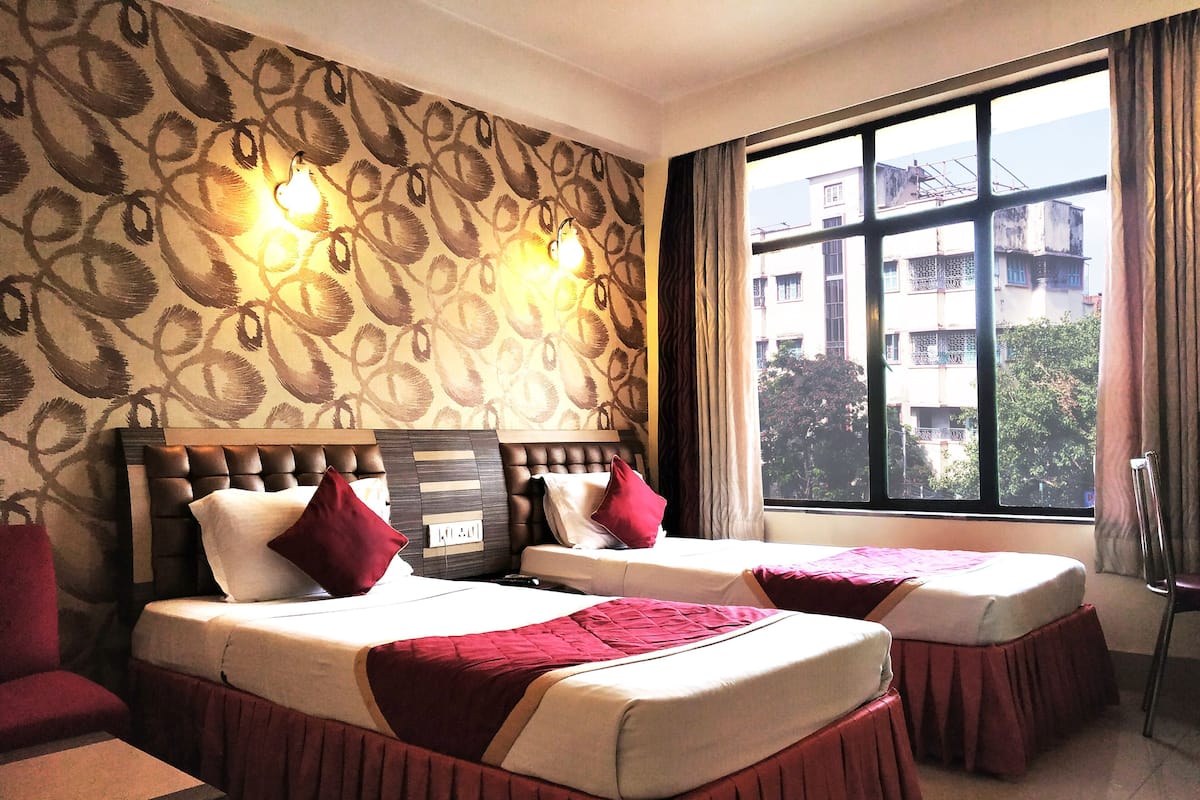 A hotel room with two beds, a patterned wall, a window with a view of buildings, and a chair.