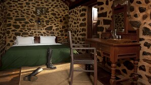 A rustic room with a stone wall, a wooden bed with green bedding, a wooden chair, a wooden dresser, and a mirror. - Elephant Valley (Kodaikanal)