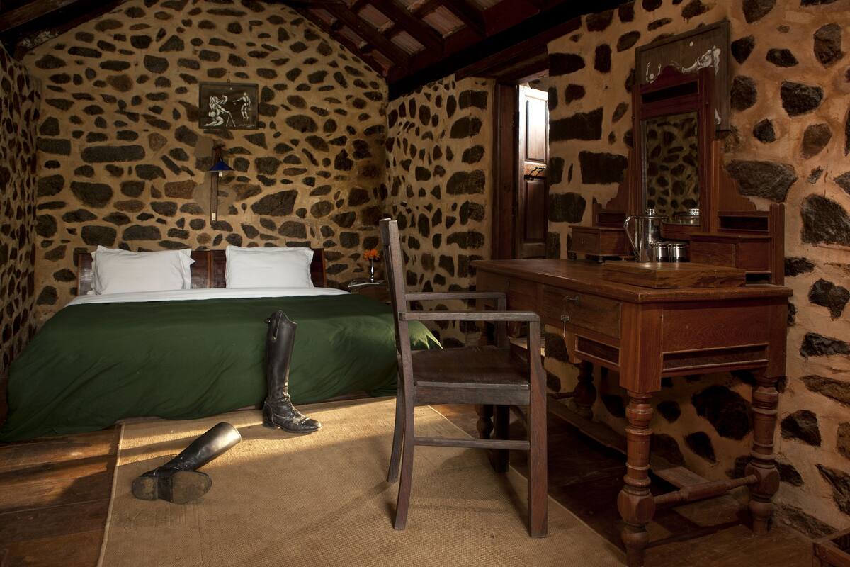 Una habitación rústica con pared de piedra, una cama de madera con ropa de cama verde, una silla de madera, un aparador de madera y un espejo.