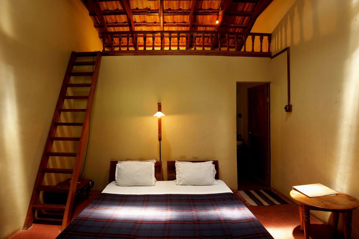 Una habitación pequeña con una cama, una escalera de madera, una mesita de noche y una puerta que da a otra habitación.