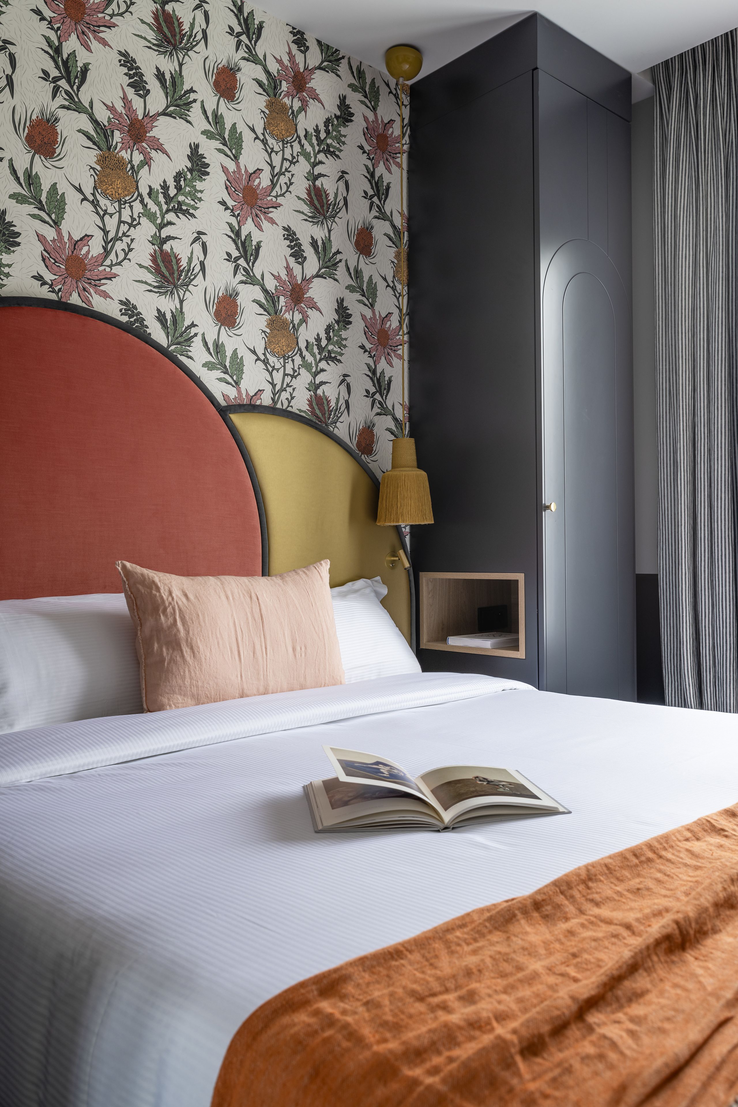 A neatly made bed with a red headboard, a bedside lamp, and a floral wallpaper pattern.