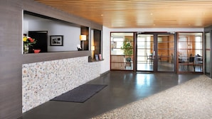 A reception area with a wooden reception desk, a pebbled floor, and a glass door leading to a dining area. - Séjours & Affaires Gex Les Rives du Leman (Gex)