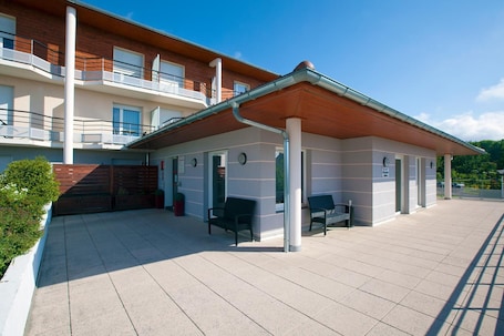 A covered outdoor area with a wooden roof, benches, and a building with balconies and windows.