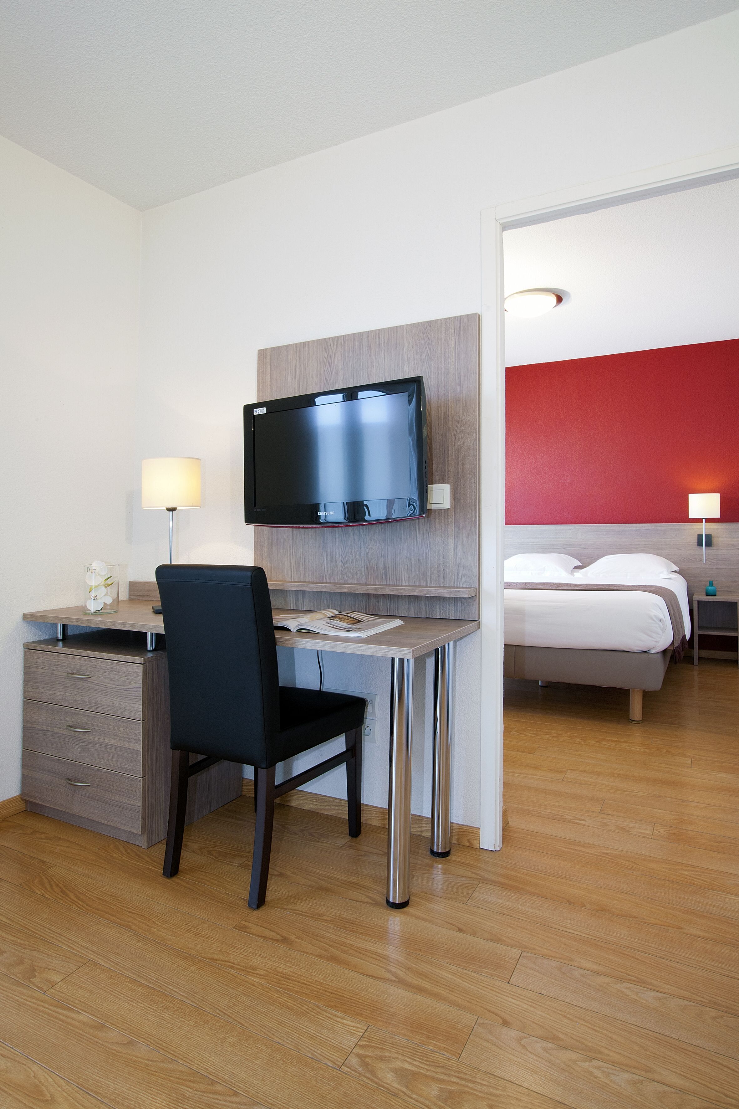 Una camera d'hotel con una scrivania in legno, una sedia nera, un letto con biancheria bianca e un televisore montato a parete.
