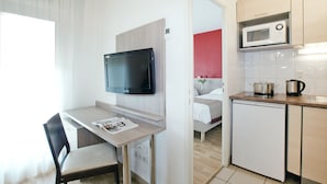 A compact hotel room with a kitchenette, a TV mounted on the wall, a small dining table, and a bed with a red accent wall. - Séjours & Affaires Gex Les Rives du Leman (Gex)