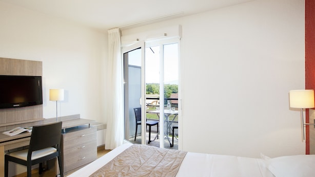 A hotel room with a bed, a desk, a chair, a television, and a balcony with a table and chairs. - Séjours & Affaires Gex Les Rives du Leman (Gex)