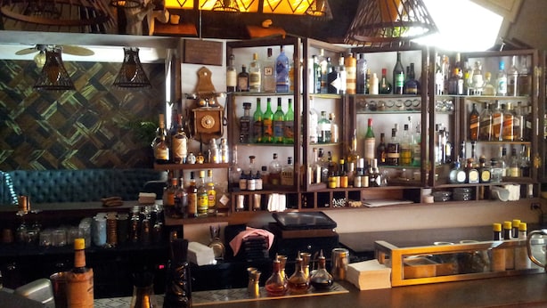 Un bar avec des étagères remplies de bouteilles d’alcool, un coin salon bleu avec des sièges rembourrés et des luminaires suspendus.