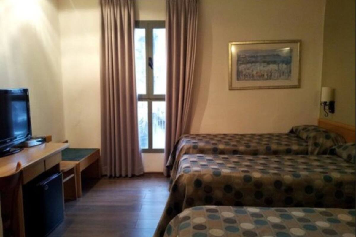 Habitación de hotel con dos camas, un escritorio con televisor, una ventana con cortinas y un cuadro en la pared.