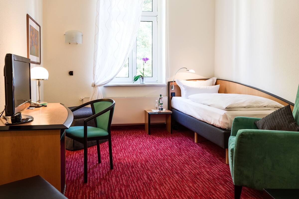 Habitación de hotel con escritorio, silla, cama y una mesita de noche con lámpara.