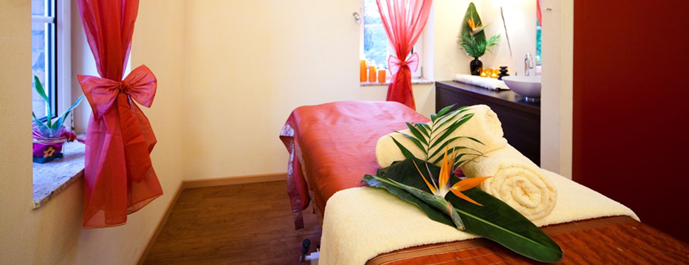Ein Wellnessraum mit Massagetisch, Handtuch mit Pflanzenmotiv und einem Fenster mit roten Vorhängen.
