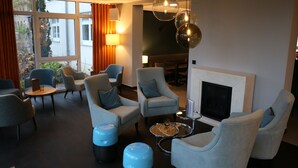 A modern lounge area with comfortable seating, a fireplace, and large windows offering a view of the outdoors. - VITALHOTEL ambiente (Bad Wilsnack)