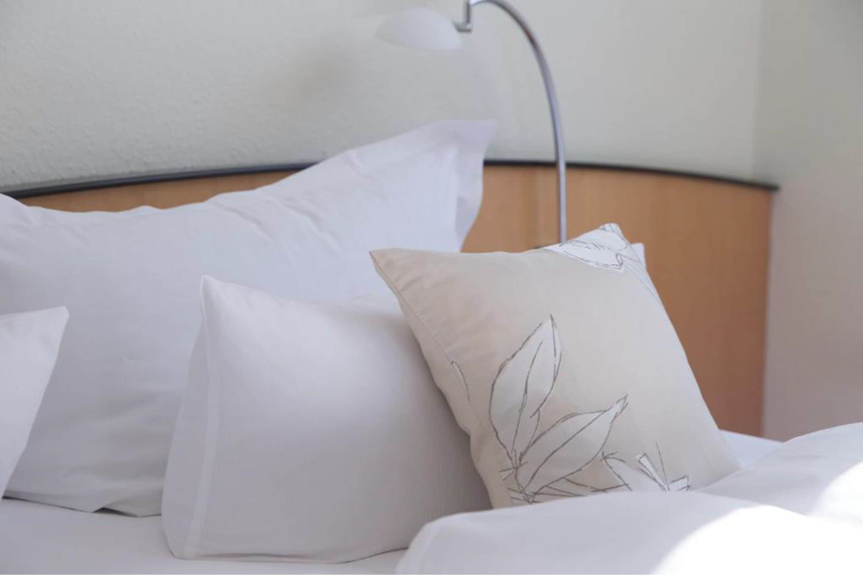 Ein ordentlich bezogenes Bett mit weißer Bettwäsche und einem einzelnen dekorativen Kopfkissen mit Blattform-Motiv.
