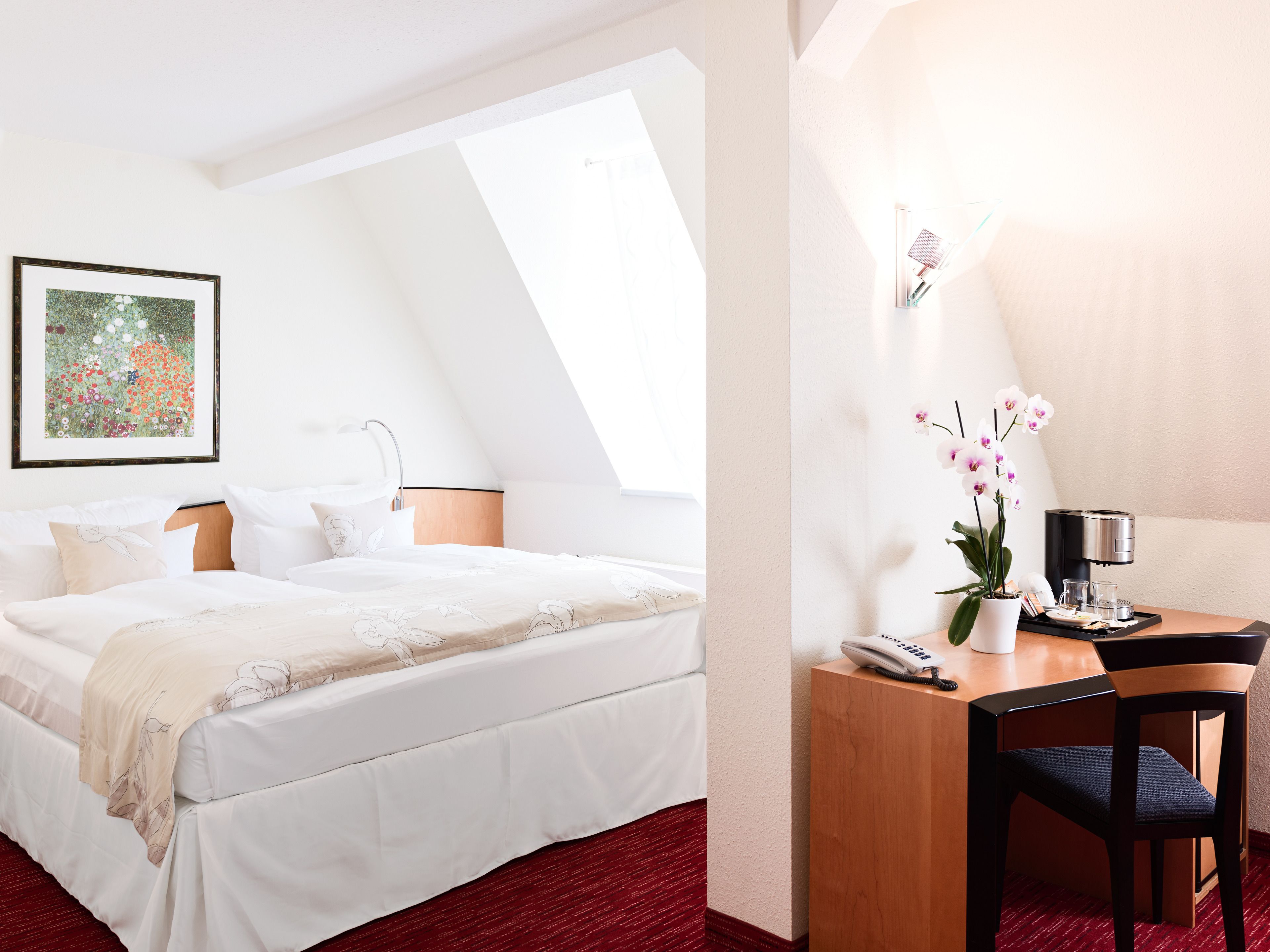 Habitación de hotel con cama, escritorio con silla, teléfono, cafetera y una orquídea en maceta.