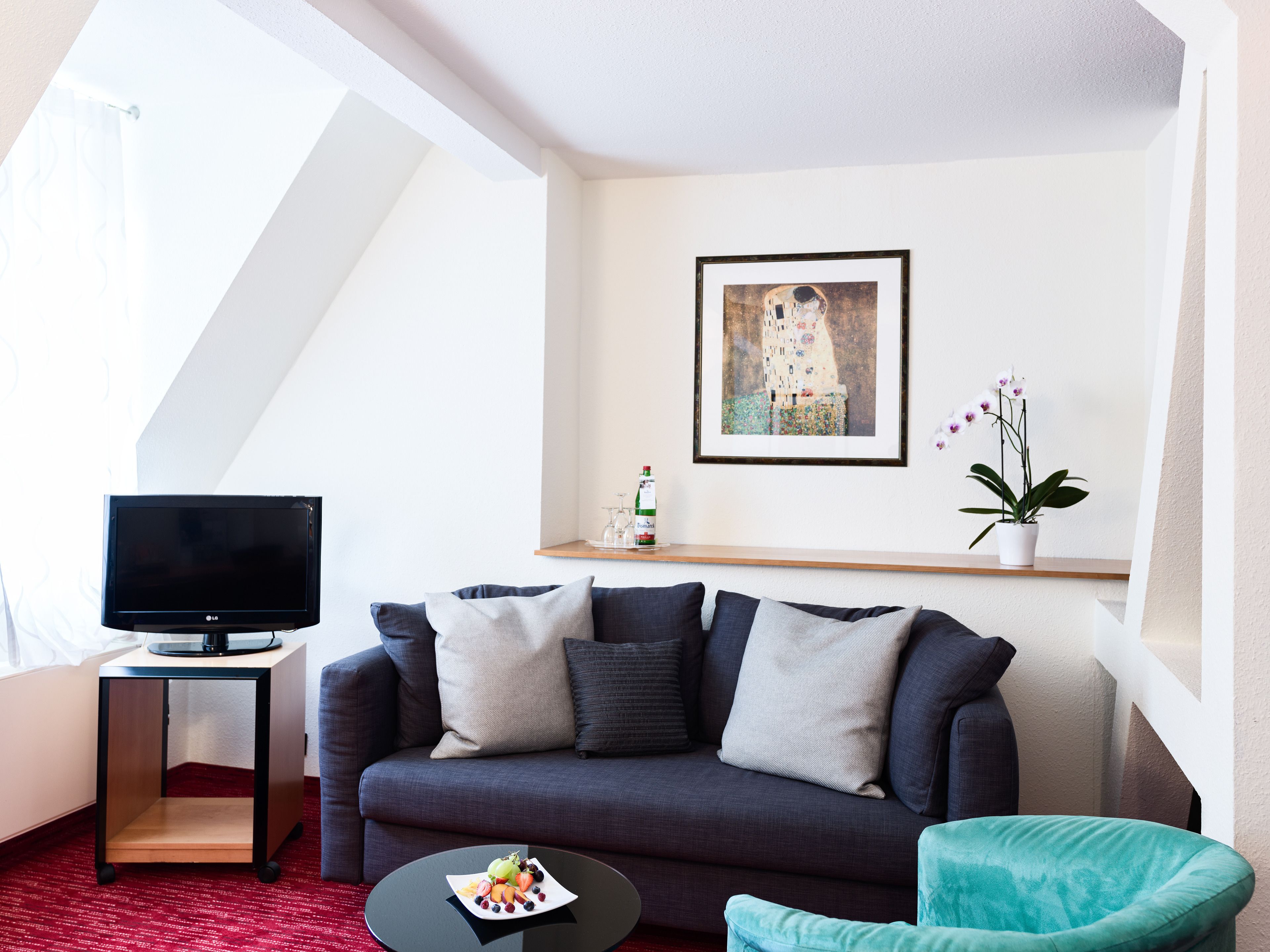 A modern living room with a dark sofa, a small table with fruit, a flat-screen TV, a framed artwork, and a potted plant.