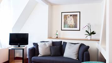A modern living room with a dark sofa, a small table with fruit, a flat-screen TV, a framed artwork, and a potted plant.