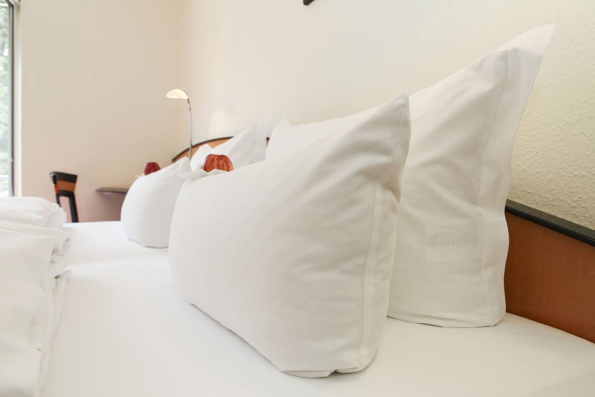 Una cama bien hecha con sábanas y almohadas blancas, una mesita de noche con lámpara, y una silla en una habitación de hotel.
