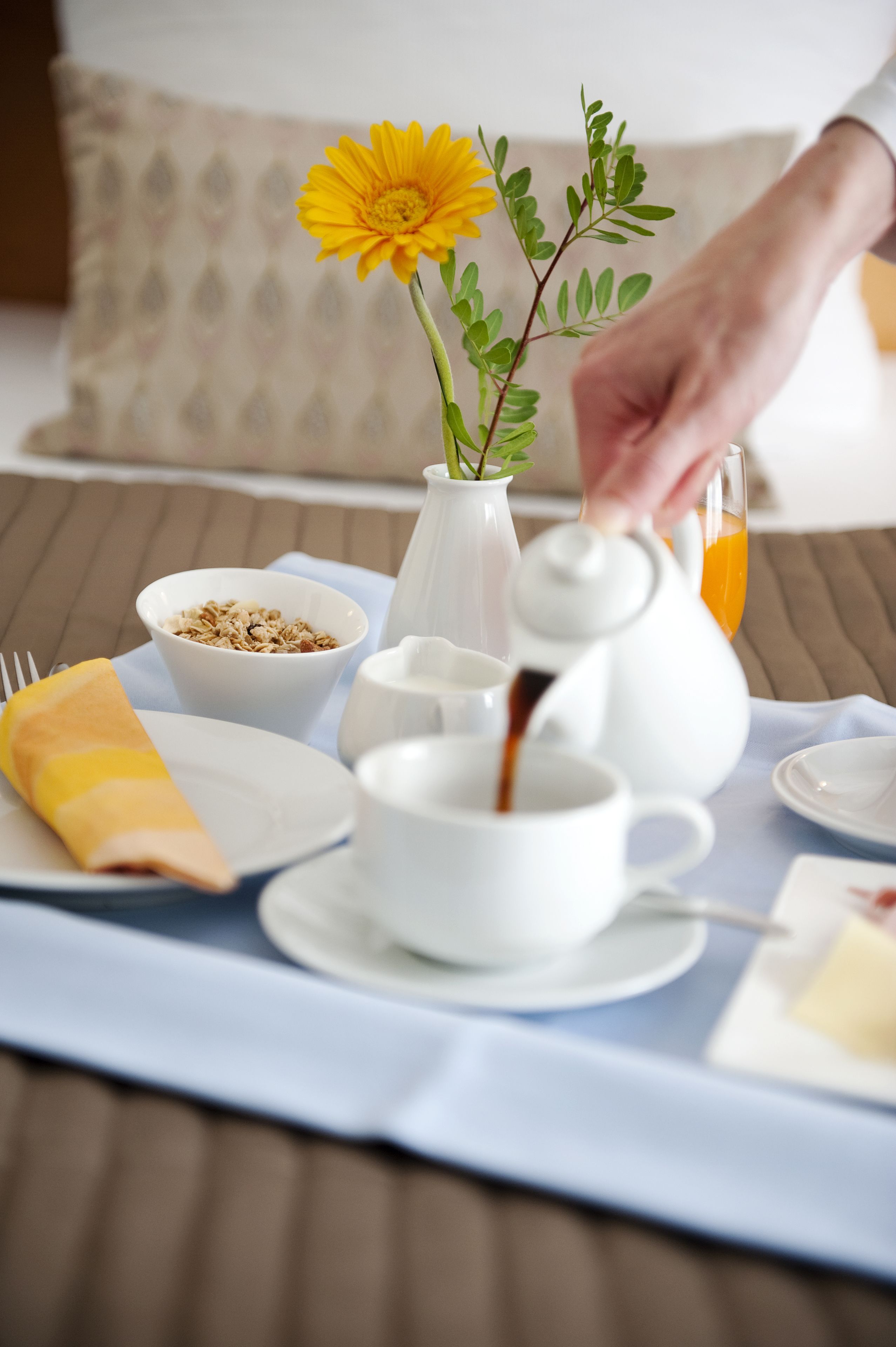 A breakfast setup with a teapot, cups, a bowl of cereal, a vase with a flower, and a napkin on a table.