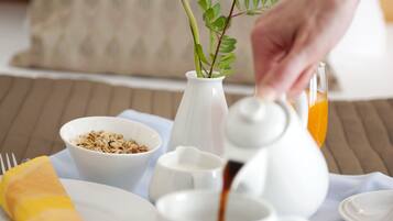 A breakfast setup with a teapot, cups, a bowl of cereal, a vase with a flower, and a napkin on a table.