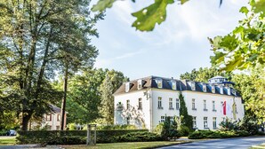 A large, two-story building with a white facade and dark roof, surrounded by lush greenery and a clear sky. - VITALHOTEL ambiente (Bad Wilsnack)