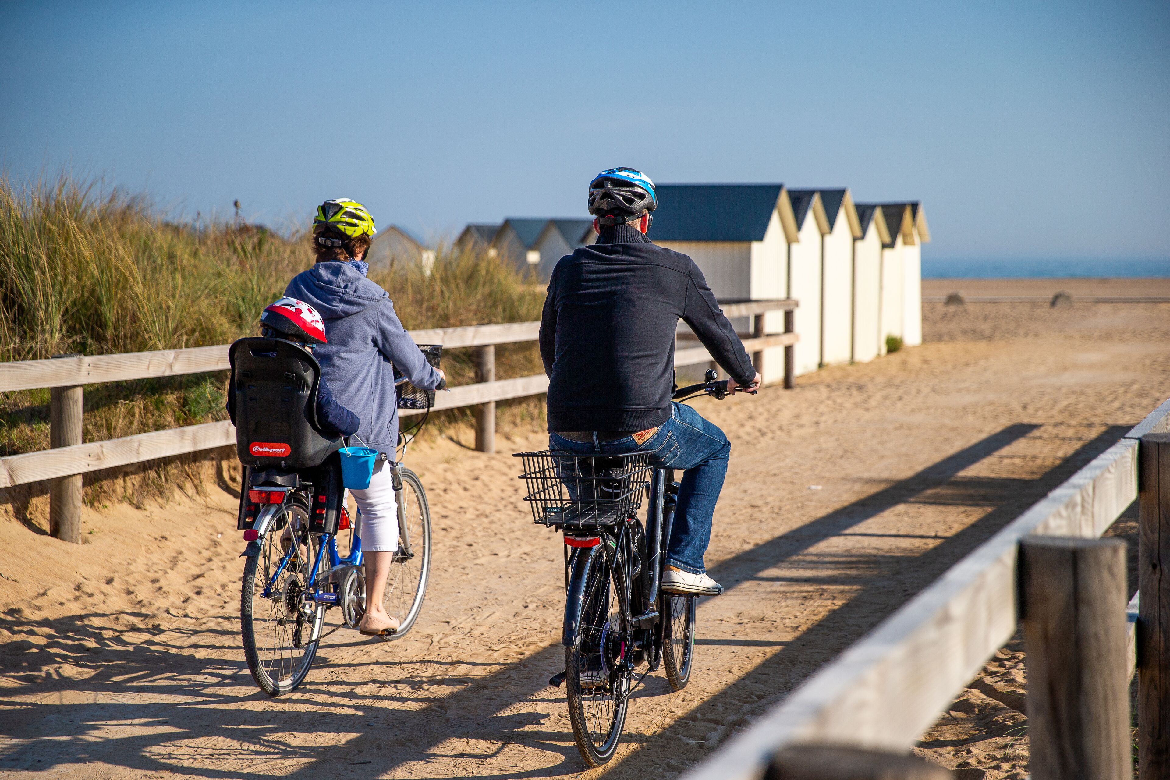 Two people on bicycles on a sandy path with wooden fences and beach huts in the background.
