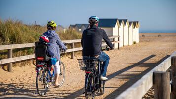 Two people on bicycles on a sandy path with wooden fences and beach huts in the background.