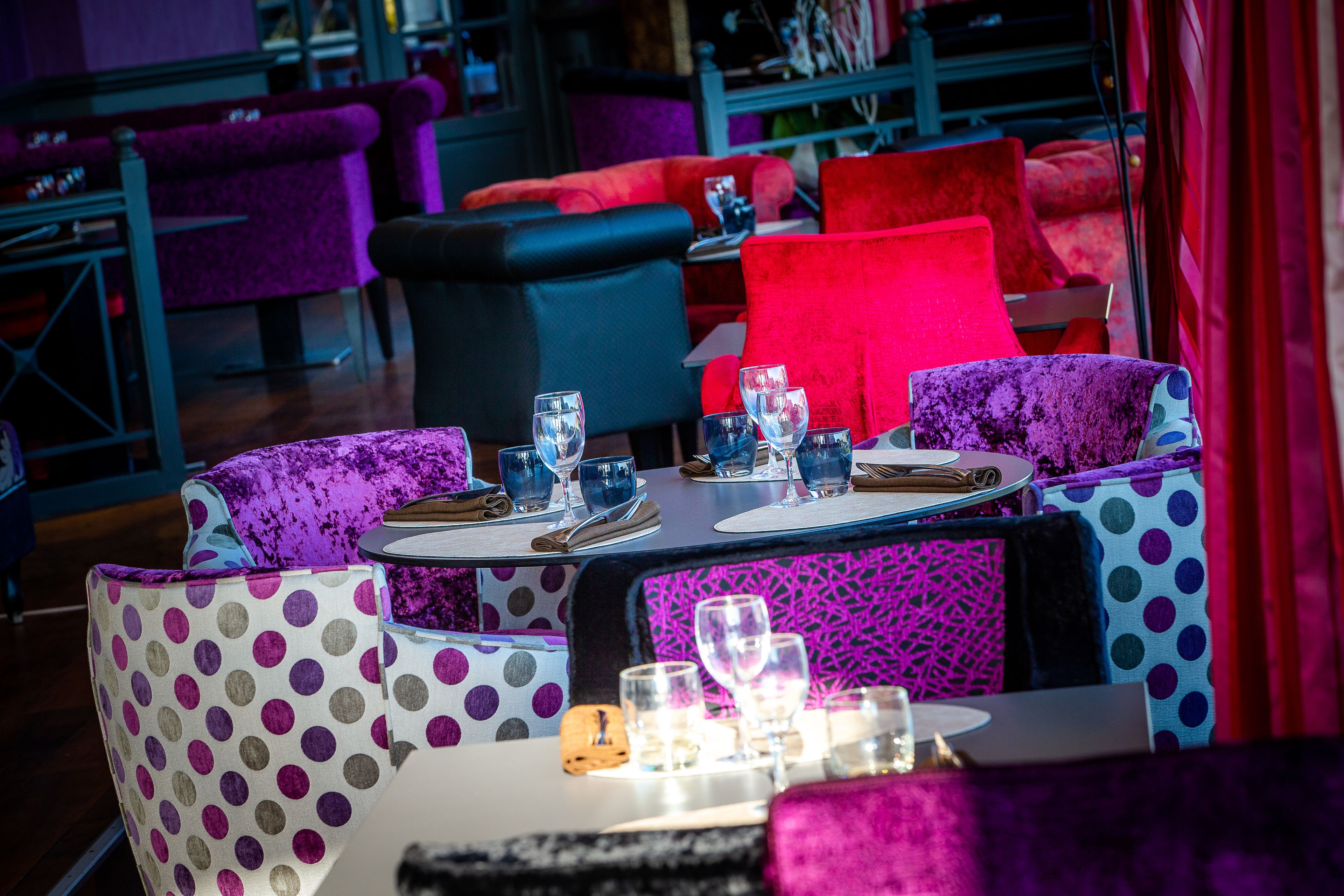 L’intérieur d’un restaurant, avec des chaises aux couleurs vives, violettes et rouges, des tables dressées avec des verres et des couverts, et un sol à motifs.