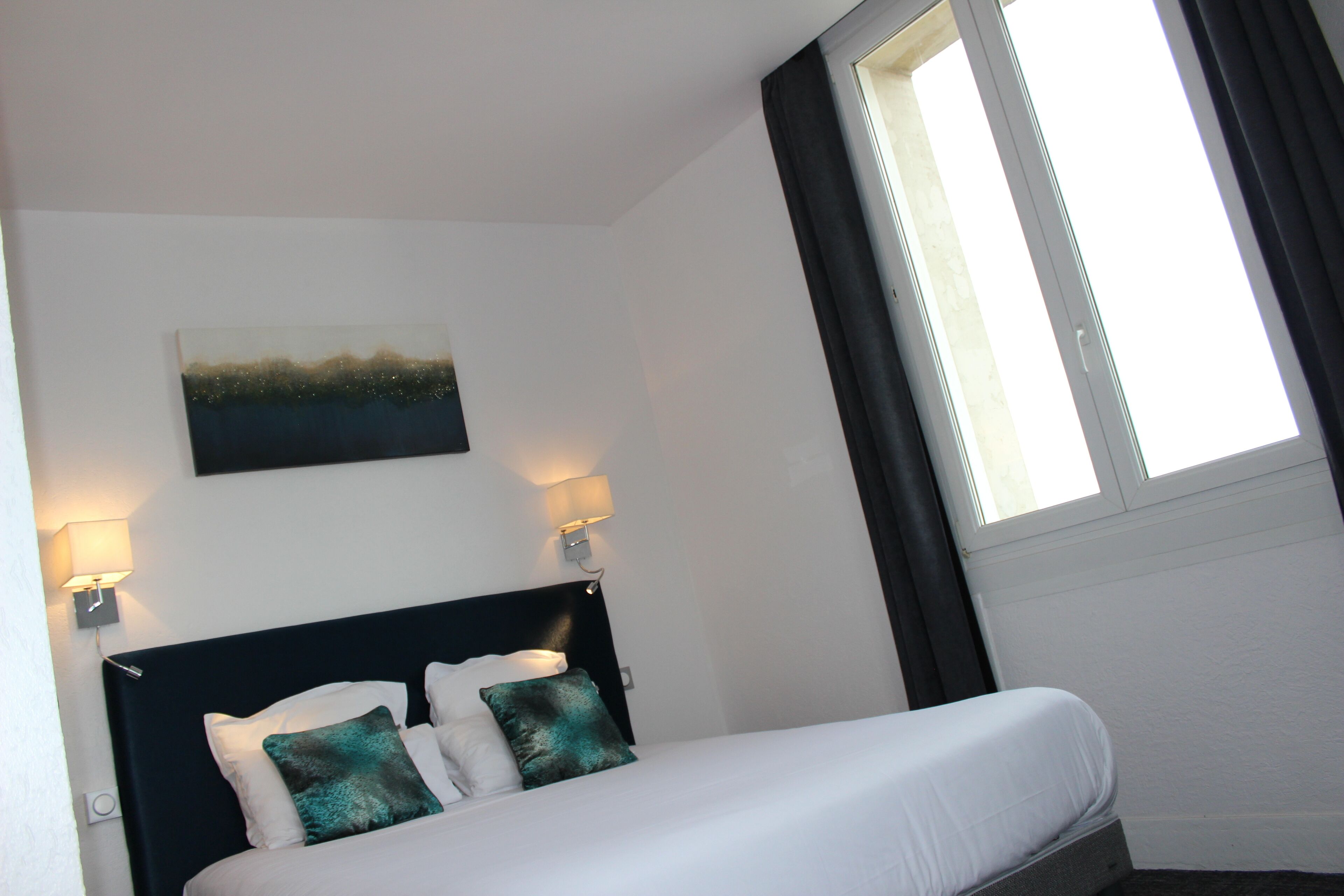 Een hotelkamer met een bed, twee wandlampen, een schilderij en een raam met gordijnen.
