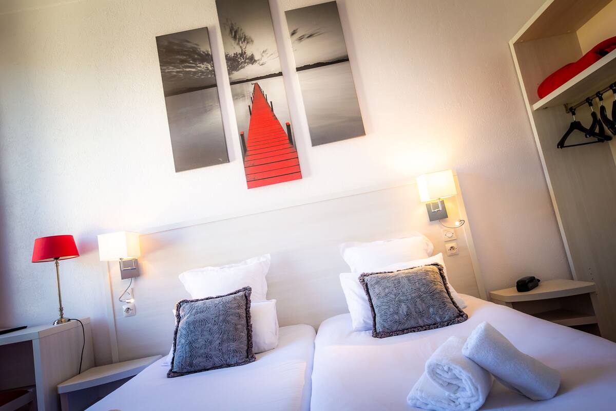 A hotel room with a bed, two bedside tables, a red lamp, wall-mounted lights, and a red decorative piece above the bed.