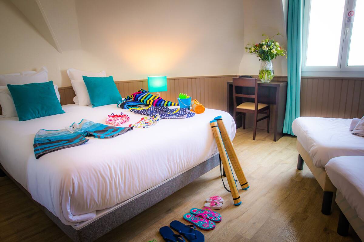 A hotel room with a large bed, a smaller bed, a desk, a chair, a lamp, a vase with flowers, and a window with curtains.