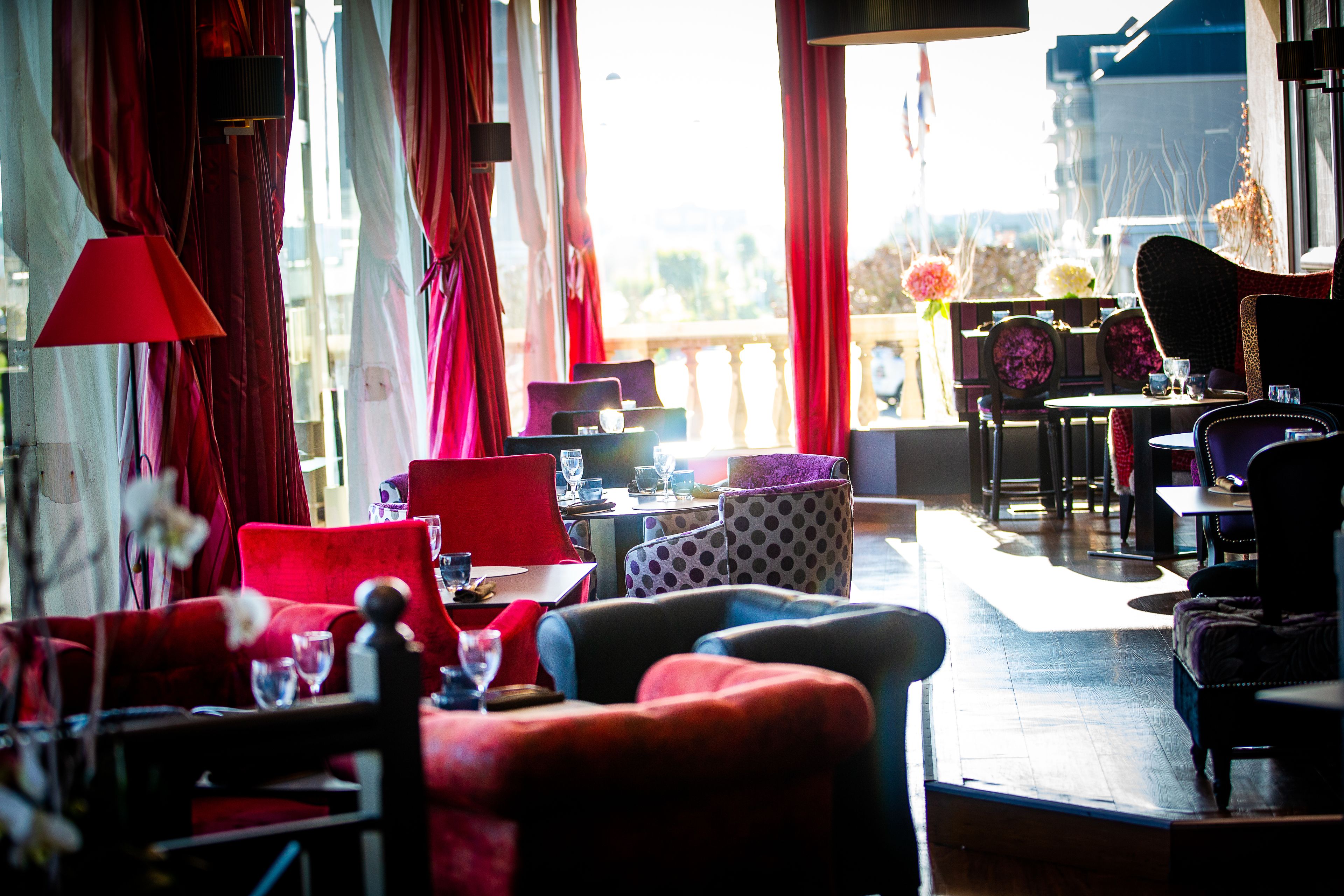 A well-lit room with red curtains, a variety of seating options including sofas and chairs, and a view of the outdoors through large windows.