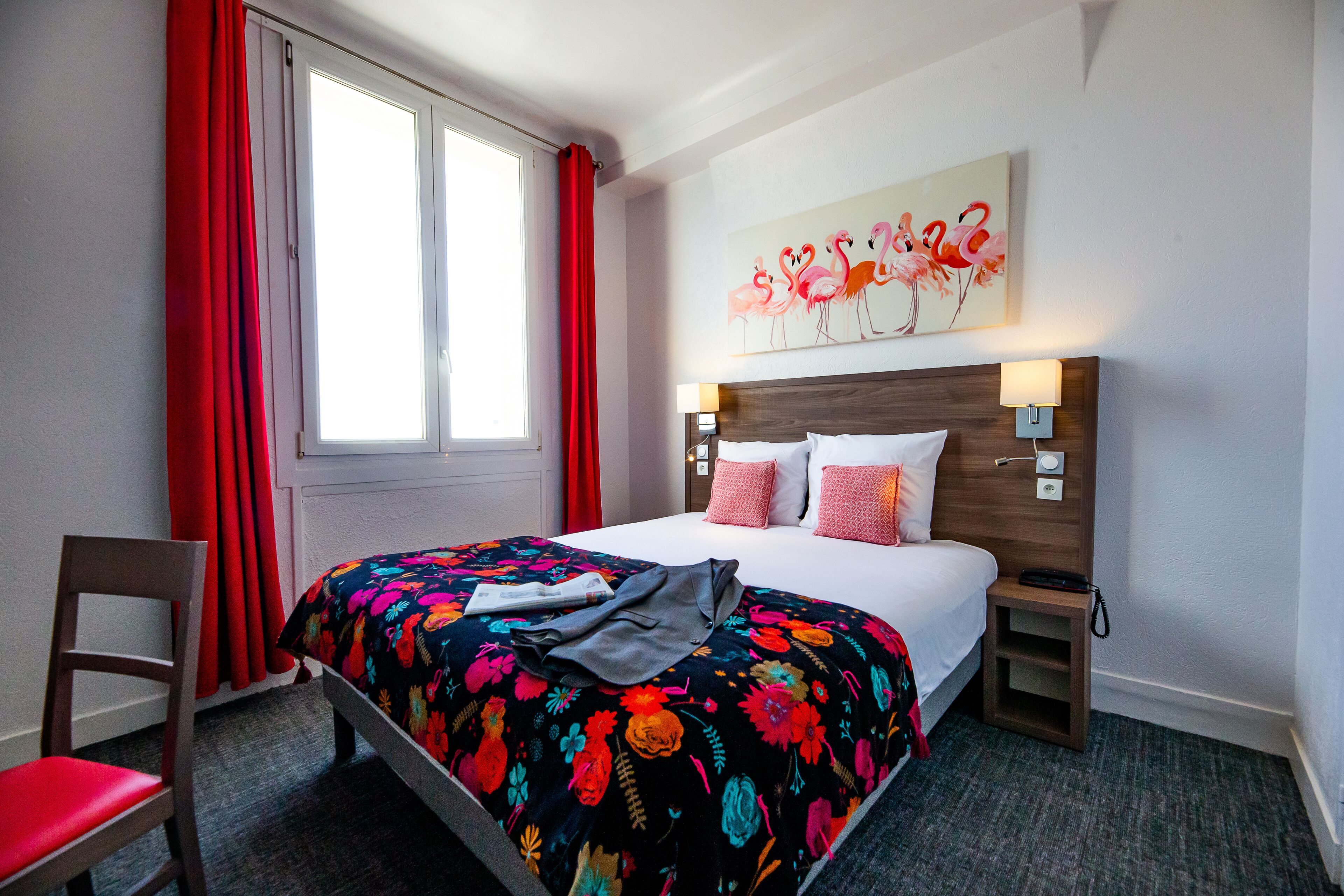 Habitación de hotel con cama, una silla, una mesita de noche, una lámpara y un cuadro de flamencos.