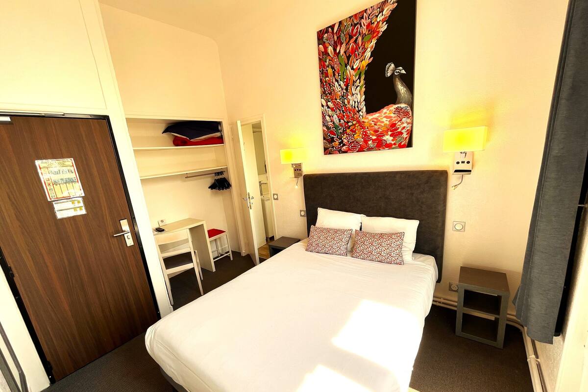 A hotel room with a bed, a desk, a chair, a wardrobe, and a painting on the wall.
