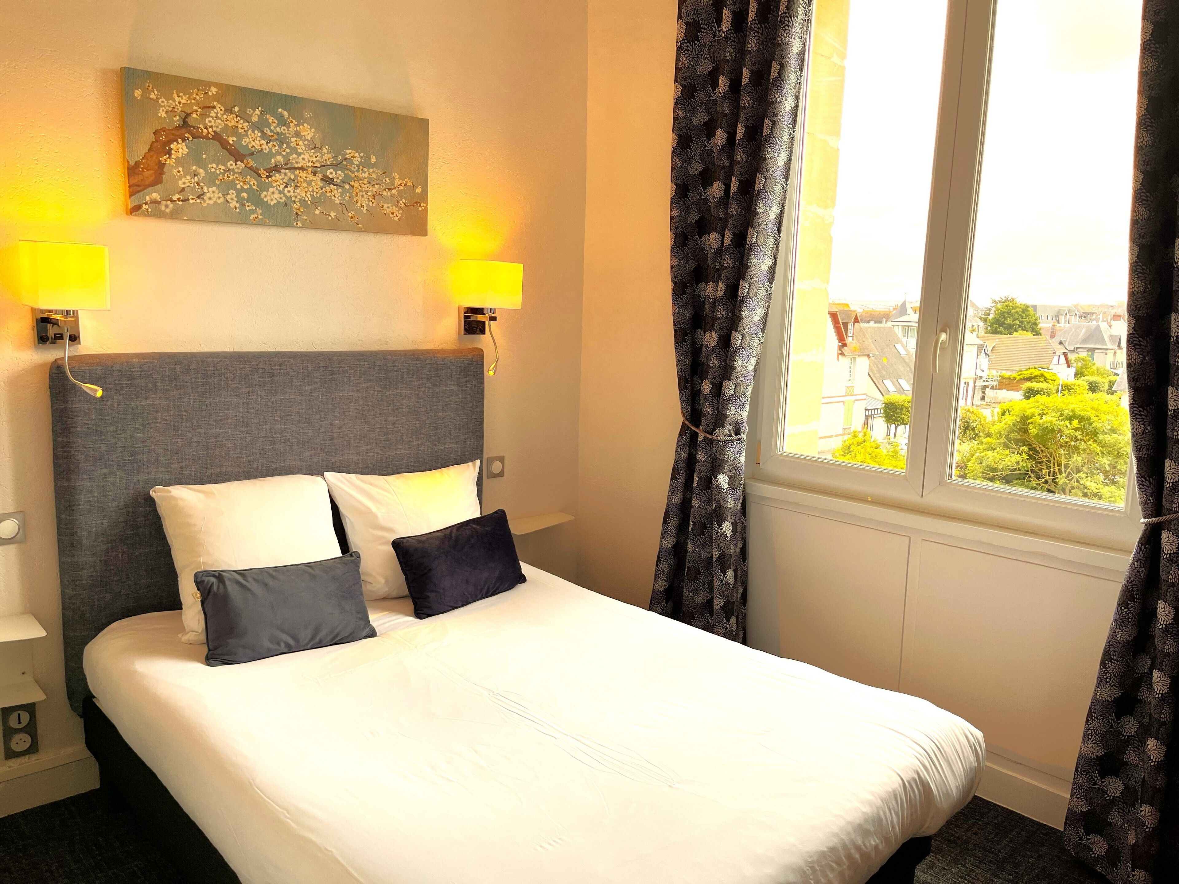 A hotel room with a bed, two wall-mounted lamps, a painting, and a window with curtains.