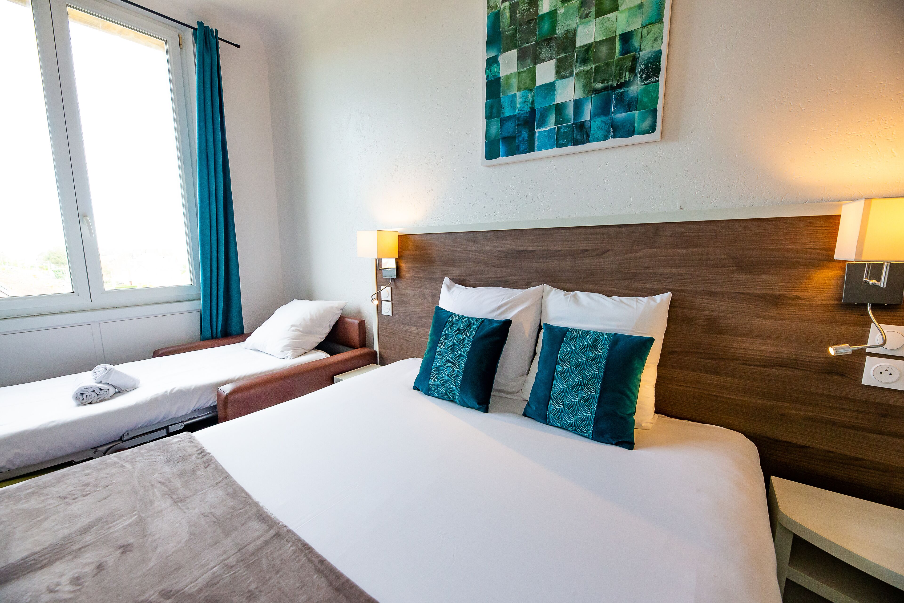 Een hotelkamer met een groot bed, twee kleinere bedden, een raam met gordijnen, een kunstwerk aan de muur en bedlampjes.