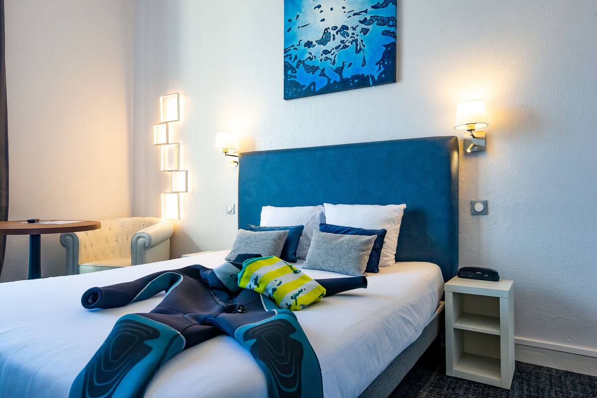 A hotel room with a bed, a chair, a small table, and a nightstand with a lamp.