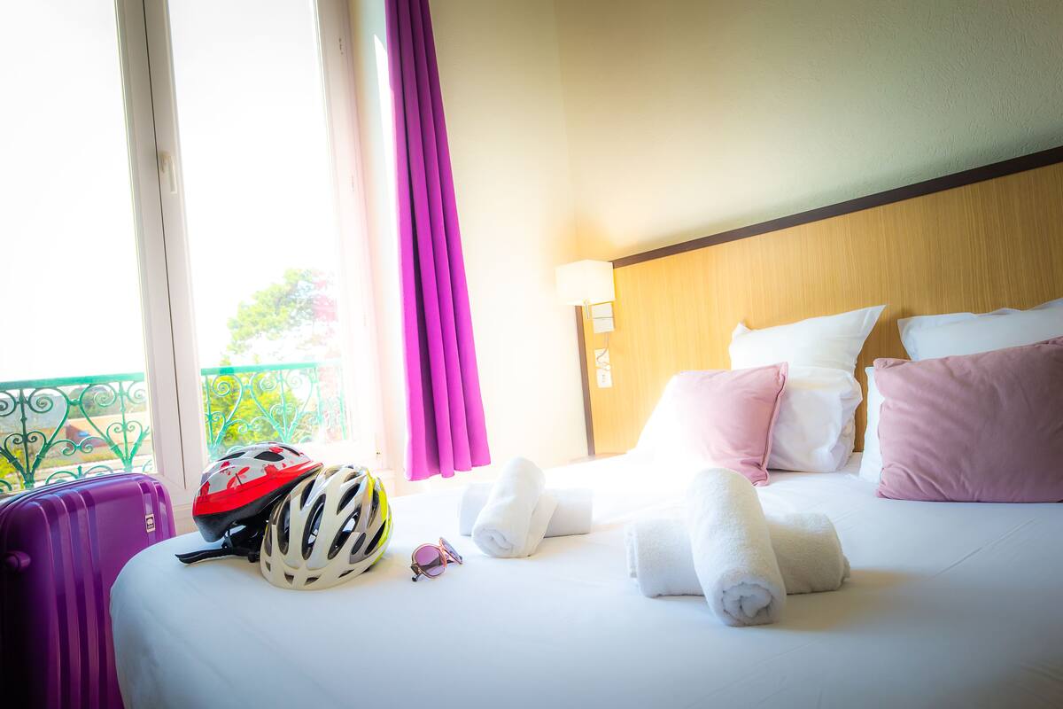 A hotel room with a bed, pillows, a headboard, a window with curtains, a suitcase, and a helmet.