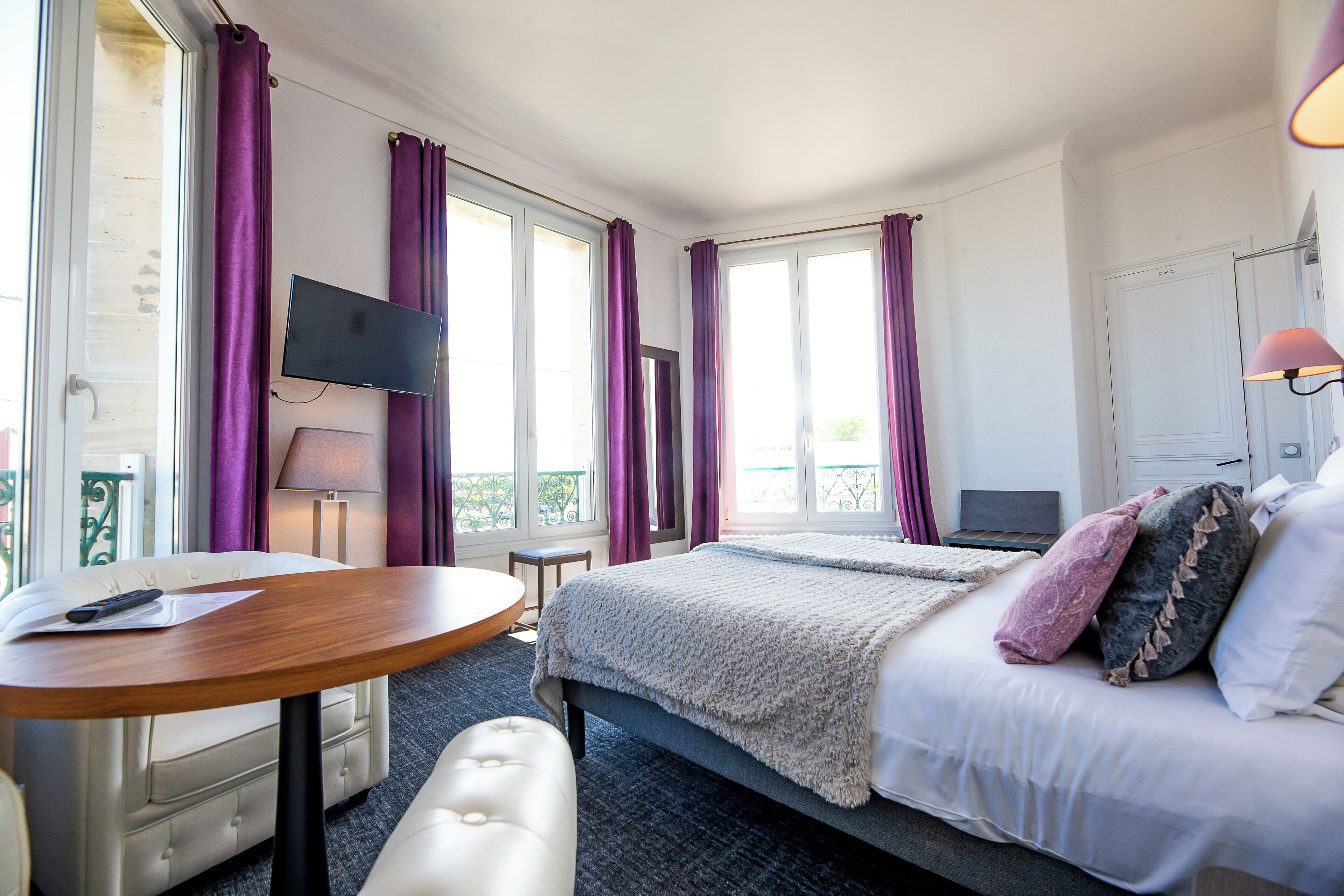 A hotel room with a bed, a desk with a chair, a TV, and large windows with purple curtains.