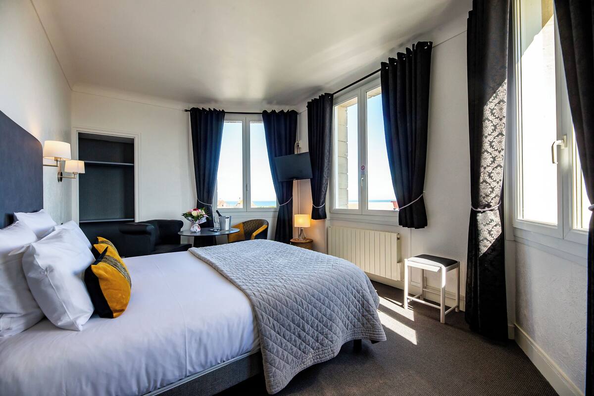 A hotel room with a bed, a desk, a chair, a lamp, and a view of the outdoors.