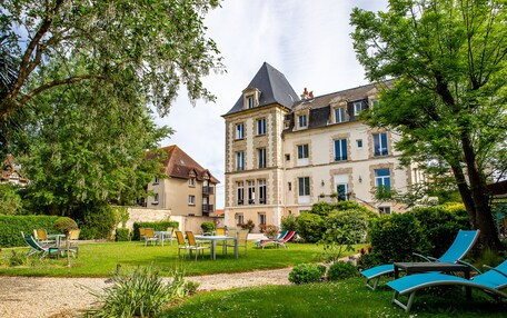 A large, historic building with a prominent tower and multiple windows. There is a well-maintained garden with a gravel path, chairs, and tables.