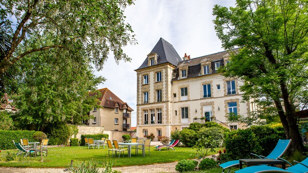 A large, historic building with a prominent tower and multiple windows. There is a well-maintained garden with a gravel path, chairs, and tables. - La Villa Andry (Ouistreham)