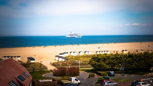 A beach view with a cruise ship in the distance, beach huts, and a parking area with cars. - La Villa Andry (Ouistreham)