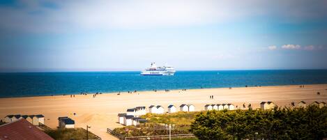 Vista de una playa con un crucero a lo lejos, cabañas de playa y un estacionamiento con coches.