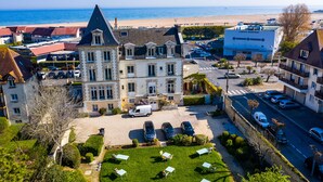 A large building with a prominent tower, a parking area with several cars, and a view of the beach in the background. - La Villa Andry (Ouistreham)