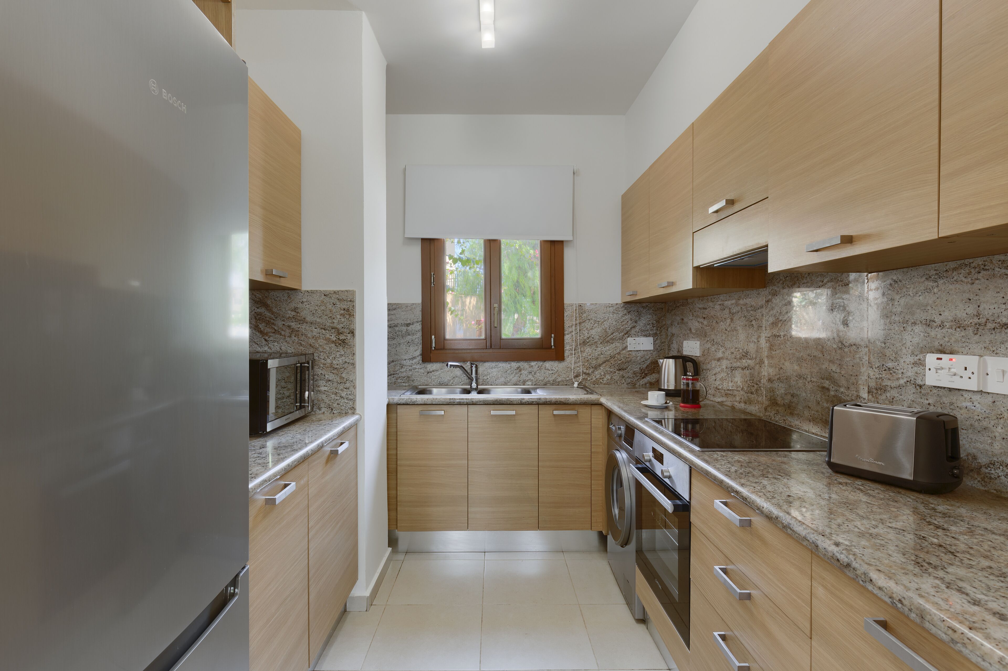 A modern kitchen with wooden cabinets, a stainless steel refrigerator, a microwave, a toaster, and a window above the sink.