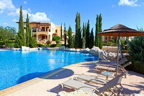 A swimming pool with lounge chairs and umbrellas, surrounded by tall trees and a building with balconies.