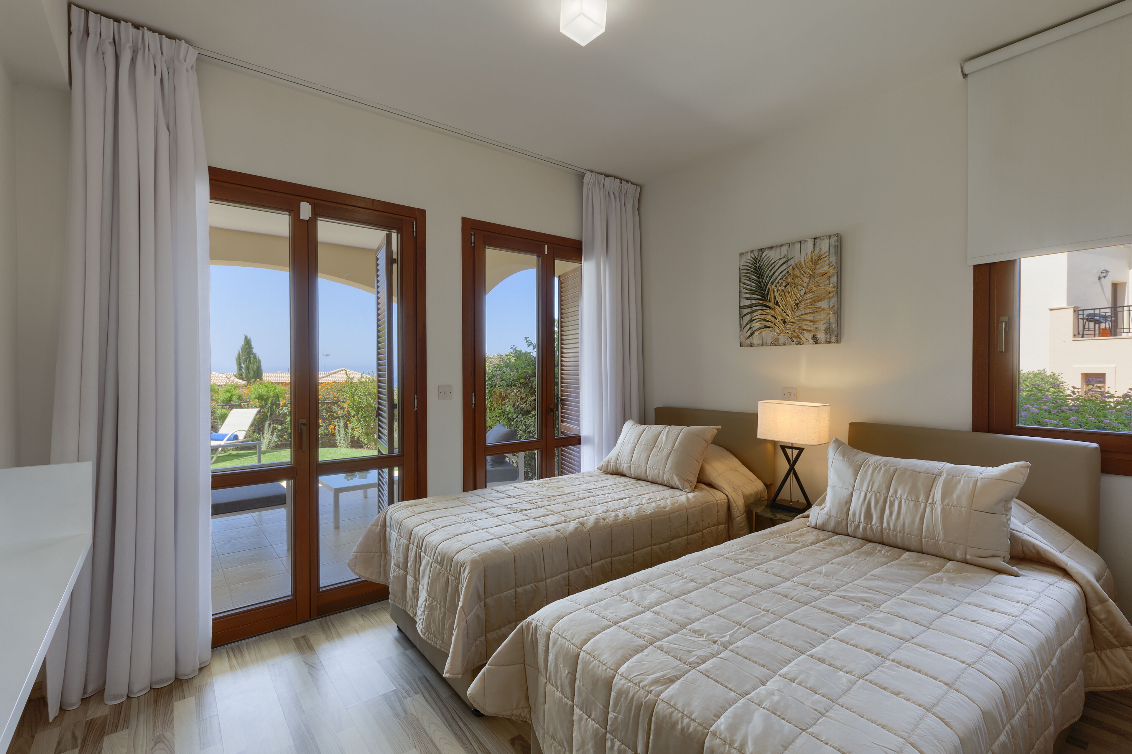 A hotel room with two beds, a window with curtains, a lamp, and a painting on the wall.